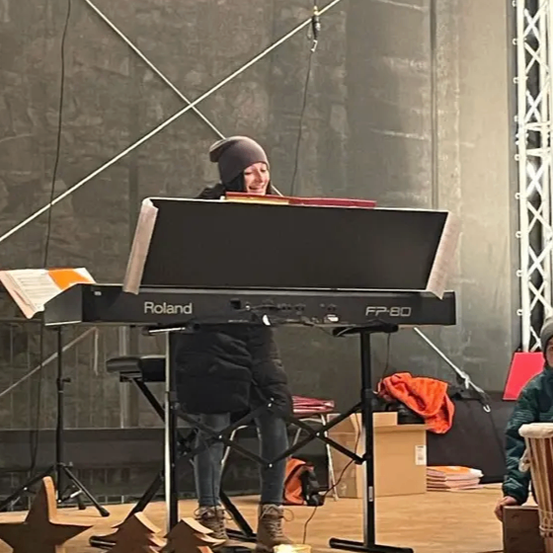 Eine Person steht auf einer Bühne und spielt eine Roland-Tastatur. Sie trägt einen Hut und Stiefel. Es gibt Kerzen, ein Schlagzeug und Weihnachtsbäume um sie herum.