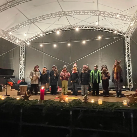 Eine Gruppe von Kindern steht auf der Bühne, gekleidet in Winterkleidung, hält Kerzen und trägt Mützen. Sie sind von Weihnachtsdekorationen umgeben, einschließlich eines Sterns und einer Kerze. Ein Klavier ist im Hintergrund.