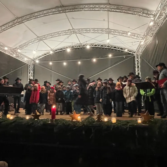 Eine Gruppe von Menschen ist auf einer Bühne unter einem Metallrahmen versammelt und singt. Kerzen und Sterne schmücken die Bühne. Einige tragen Hüte. Ein Pianist steht links.