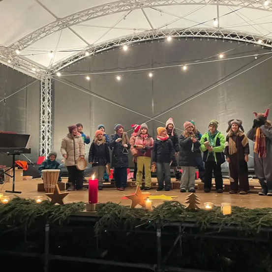 Eine Gruppe von Kindern tritt auf einer Bühne auf, geschmückt mit Weihnachtsdekorationen und Kerzen, unter einem weißen Zelt.