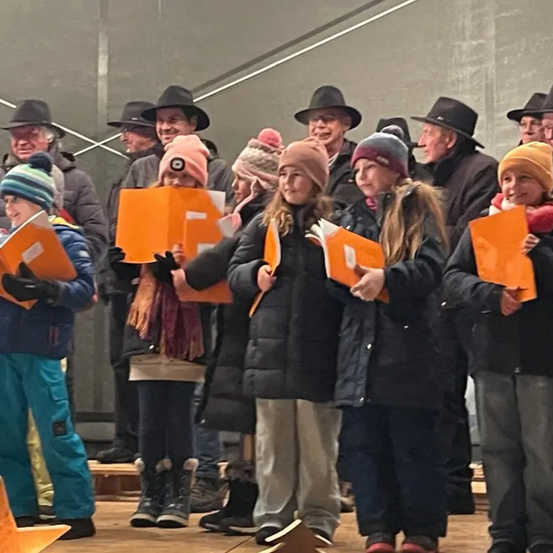 Eine Gruppe von Kindern und Erwachsenen, einige mit Mützen, stehen gemeinsam auf einer Bühne, halten orange Bücher und singen. Im Hintergrund befindet sich eine graue Wand und Treppe.