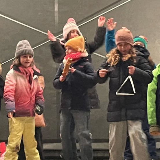 Eine Gruppe von Kindern, alle warm gekleidet, steht in einer Reihe, einige mit Musikinstrumenten. Das Mädchen links trägt eine rosa Jacke und gelbe Hosen. Das Mädchen neben ihr hält ein Dreieck. Das Mädchen in der Mitte hält eine Flöte.