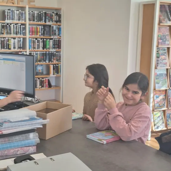 Zwei Mädchen sitzen an einem Schreibtisch in einer Bibliothek. Eines hält Bücher, und die andere lächelt, beide scheinen glücklich zu sein. Hinter ihnen steht ein Bücherregal voller Bücher. Ein Computermonitor, eine Box und Akten sind auf dem Schreibtisch.