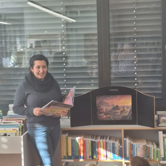 Eine Frau liest Kindern in einer Bibliothek ein Buch vor. Die Kinder sitzen vor ihr auf dem Boden.