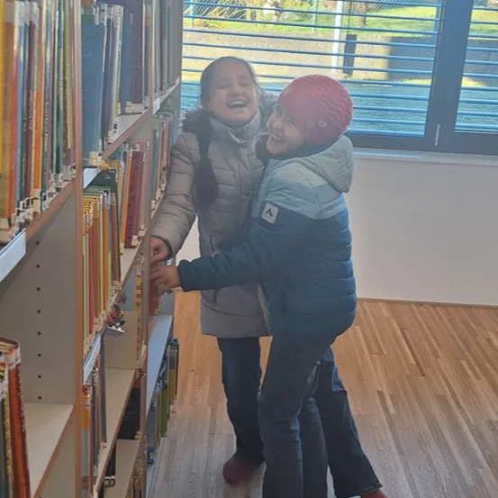 Zwei Kinder stehen in einer Bibliothek. Sie schauen auf Bücher in den Regalen. Sie lächeln.