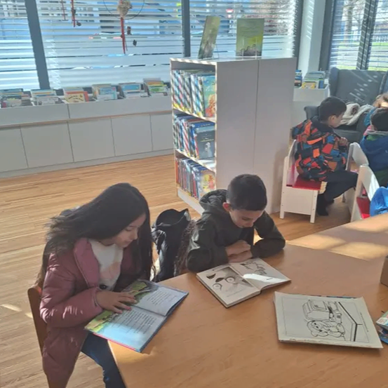 Zwei Kinder sitzen an einem Tisch in einer Bibliothek und lesen Bücher. In der Nähe sitzen weitere Kinder auf Stühlen und lesen. Die Bibliothek hat Holzböden und Bücherregale, die mit Büchern gefüllt sind.