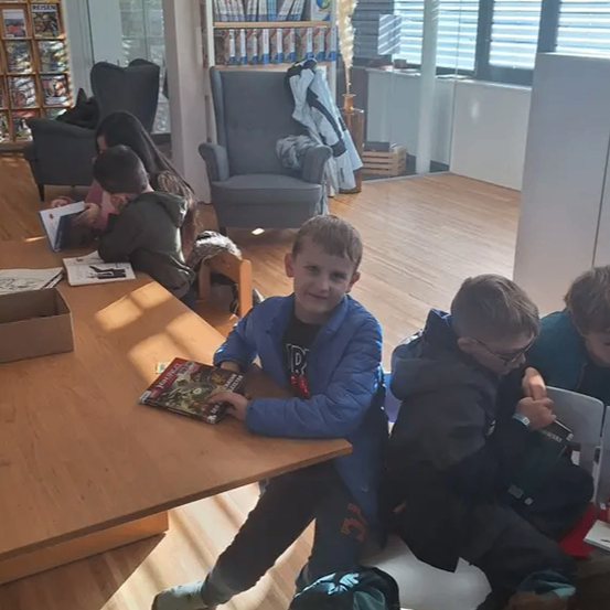 In einem Raum mit Holzböden sitzen mehrere Kinder um einen Tisch herum und lesen Bücher. Ein Junge in einer blauen Jacke sitzt mit einem Buch in der Hand am Tisch.