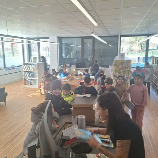 In einer Bibliothek sitzen Kinder an Schreibtischen und eine Frau entleiht Bücher. Einige Kinder stehen hinter dem Schreibtisch und schauen sich Bücher an.