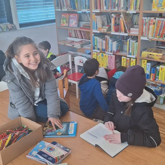 Eine Gruppe von Kindern in einer Bibliothek, wobei ein Mädchen lächelt und ein Buch ansieht und ein anderes ein Buch auf dem Tisch liest.