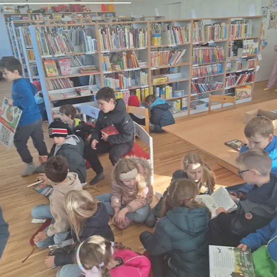 Eine Gruppe von Kindern sitzt auf dem Boden in einer Bibliothek, jeder in sein eigenes Buch vertieft. Sie sitzen im Halbkreis, einige mit Stirnbändern, während Regale voller Bücher sie umgeben.