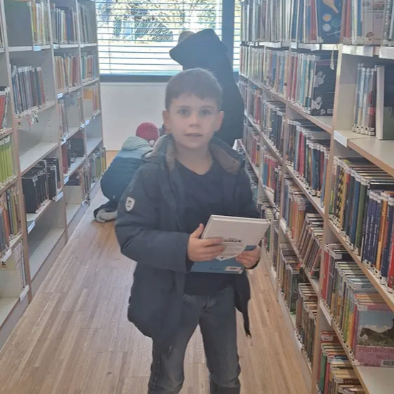 Ein Junge steht in einer Bibliothek, ein Buch in der Hand, mit Regalen voller Bücher dahinter und einer Person, die auf dem Boden sitzt.