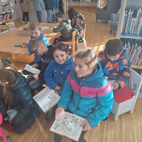 Mehrere Kinder sitzen auf dem Boden in einer Bibliothek und lesen Bücher. Einige sitzen auf Stühlen, während andere auf dem Boden sitzen.
