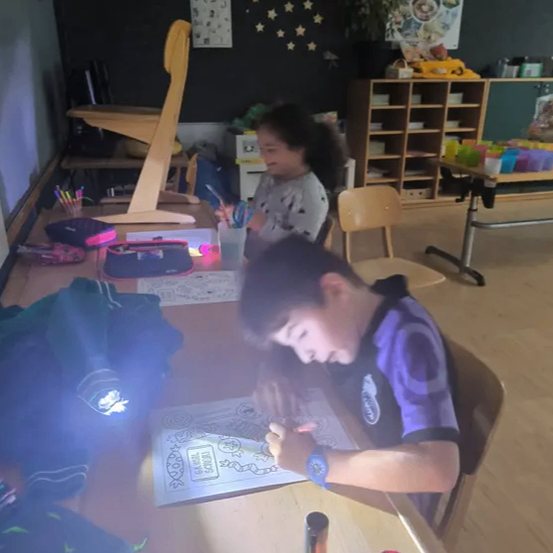 Ein Junge malt eine Seite mit einer Taschenlampe, während ein Mädchen mit Scheren neben ihm sitzt. Sie sind in einem Klassenzimmer mit Holzböden und Regalen.
