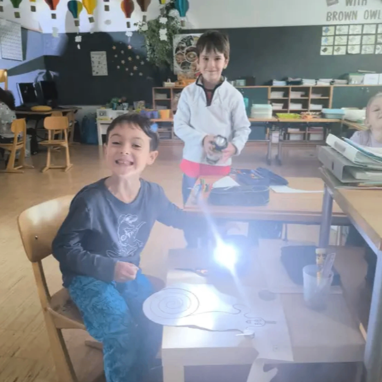 Zwei Kinder sind in einem Klassenzimmer, eines mit einer Taschenlampe und das andere mit einer Kamera. Ein Whiteboard ist hinter ihnen mit bunten Heißluftballons.