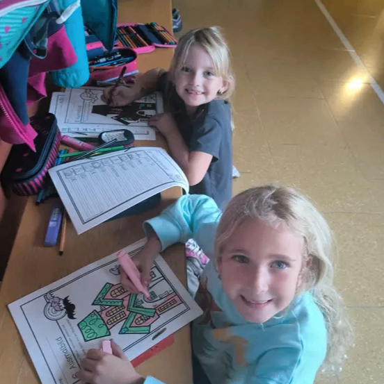 Zwei Mädchen sitzen an einem Tisch und färben ein Arbeitsblatt aus. Ein Mädchen hat einen rosa Marker und färbt ein Haus, während das andere Mädchen einen Stift hat und ein anderes Arbeitsblatt ausmalt.