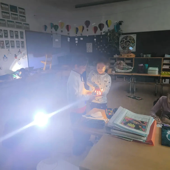 Zwei Kinder stehen in einem Klassenzimmer und halten Laternen. Ein helles Licht erhellt den Bereich. Verschiedene Gegenstände und Dekorationen sind auf den Regalen und im Raum verteilt.