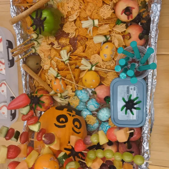 Eine Alufolieschale mit einem Halloween-Snack-Display. Es enthält verschiedene Früchte, Käsewürfel, Popcorn und einen spinnenförmigen Plastikbehälter.