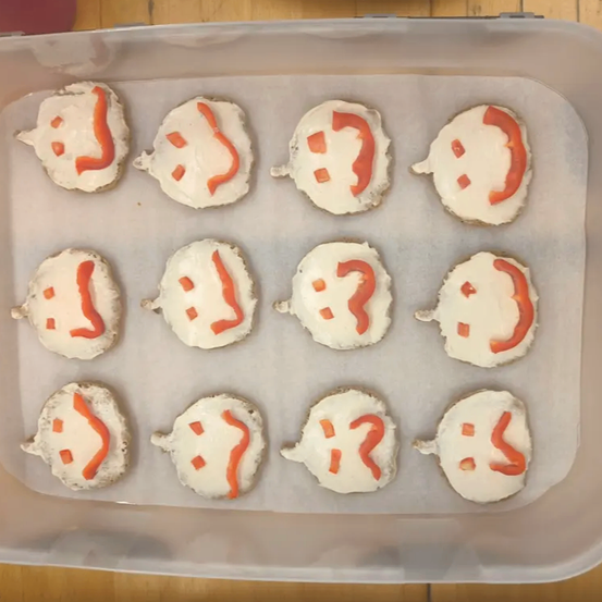 Ein Plastikbehälter enthält mehrere kreisförmige Lebensmittel, die Gesichter mit weißer Glasur und roten Fragezeichen ähneln, auf einem Backblech.