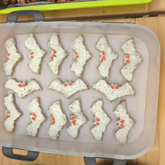 Ein Plastikbehälter enthält mehrere fledermausförmige Cracker, die jeweils mit einem roten Punkt auf einer weißen Basis gekrönt sind. Der Behälter steht auf einer Holzoberfläche.