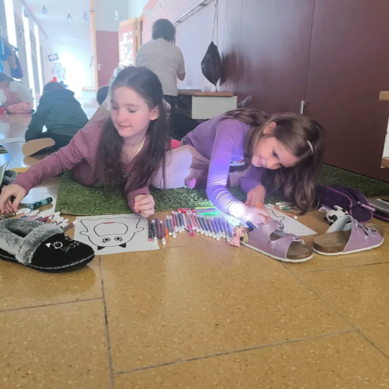 Zwei junge Mädchen färben eine Katzenzeichnung mit Buntstiften auf dem Boden aus. Sie tragen lila Schuhe.