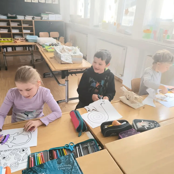 Drei Kinder sitzen in einem Klassenzimmer an Schreibtischen und sind mit Malaktivitäten beschäftigt. Ein Mädchen in einem lila Shirt malt ein Bild, während ein Junge in einem schwarzen Shirt lacht. Ein anderer Junge in einem grauen Shirt konzentriert sich auf seine eigene Kunst.