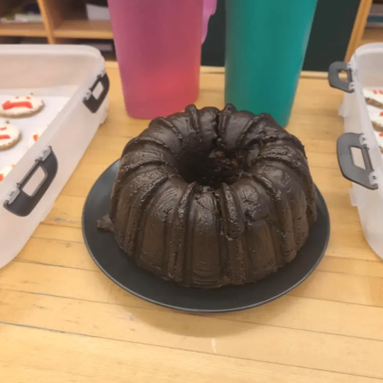 Ein Schokoladen-Gugelhupf auf einem schwarzen Teller steht auf einem Holztisch mit zwei Behältern mit Keksen und zwei farbigen Bechern im Hintergrund.