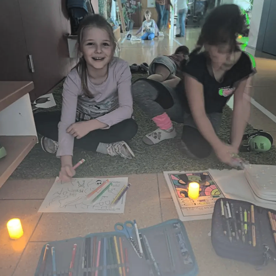 Zwei junge Mädchen sitzen auf dem Boden, malen und lächeln. Eines hält einen Stift und das andere eine Kerze. Sie sind von Malutensilien umgeben.