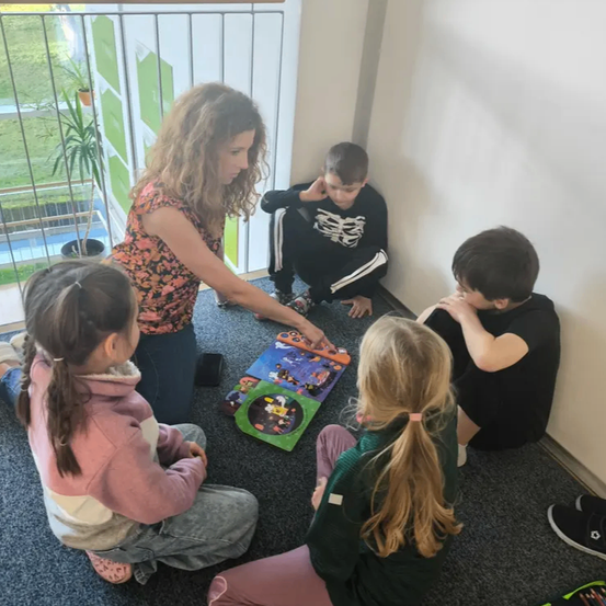 Eine Frau lehrt drei Mädchen und zwei Jungen ein Brettspiel. Sie sitzen auf dem Boden. Ein Junge bedeckt seine Ohren.