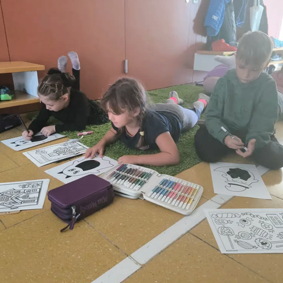 Drei Kinder malen in einem Klassenzimmer. Ein Mädchen liegt auf dem Boden mit einem Malbuch. Ein anderes Mädchen sitzt und malt. Ein Junge sitzt und hält einen Stift.
