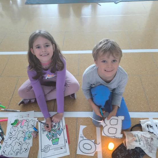 Zwei kleine Kinder sitzen auf einem gefliesten Boden und beschäftigen sich mit einer Malaktivität. Das Mädchen hält einen Marker und färbt eine Seite ein. Der Junge malt einen großen Buchstaben 'A' aus.