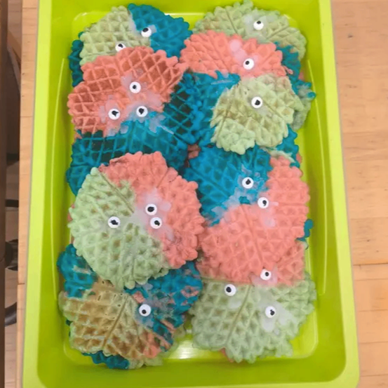 Ein grünes Tablett hält bunte Waffel-Kekse mit Augen auf einem Holztisch.