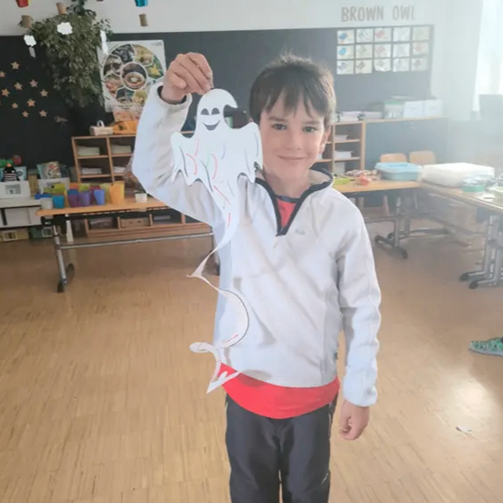 Ein junger Junge steht in einem Klassenzimmer und hält einen Papiergeist. Er trägt eine weiße Jacke und schwarze Hose. Hinter ihm befindet sich eine Tafel mit englischer Schrift. Im Hintergrund sind Tische und Stühle aufgestellt.