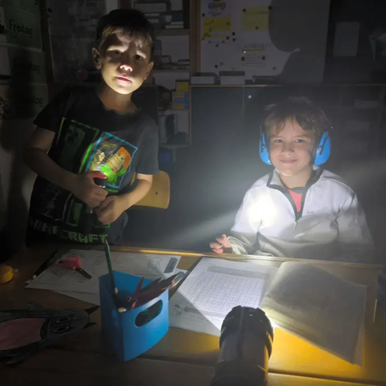 Zwei Jungen sitzen an einem Schreibtisch mit Papieren und Werkzeugen. Ein Junge hält eine Taschenlampe und der andere trägt Kopfhörer. Ein blauer Stifthalter und Scheren sind auf dem Schreibtisch.