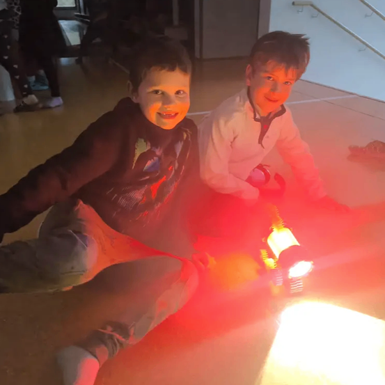 Zwei Jungen sitzen auf einem von rotem Licht beleuchteten Boden, lächeln und schauen in die Kamera. Der Junge auf der linken Seite trägt ein schwarzes Sweatshirt und orangefarbene Hose. Der Junge auf der rechten Seite trägt ein weißes Hemd und hält eine Taschenlampe.