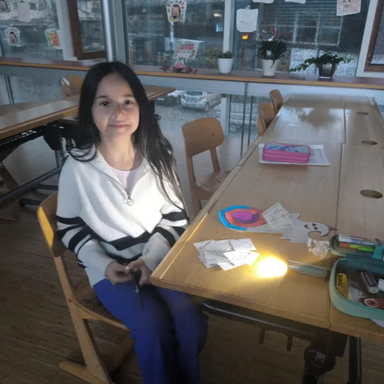 Ein junges Mädchen mit langen Haaren sitzt lächelnd an einem Schreibtisch in einem Klassenzimmer mit Fenstern und einem Fenstersims, der mit Pflanzen und Papieren geschmückt ist.