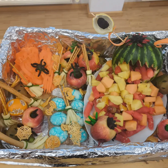 Ein Alufolientablett enthält ein Halloween-thematisches Obstplatten mit Apfelscheiben, Karotten und dekorativen Lebensmitteln, einschließlich einer geschnitzten Wassermelone und einem Apfel mit schwarzem Zentrum.