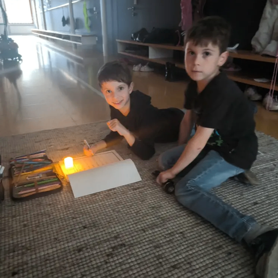 Zwei junge Jungen sitzen mit einer Kerze auf dem Boden. Ein Junge lächelt und schreibt auf ein Blatt Papier.