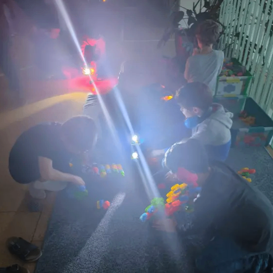 Kinder spielen mit Spielzeug auf einem Teppichboden, beleuchtet von einem hellen Licht. Zwei Kinder knien, und eines sitzt auf dem Boden.