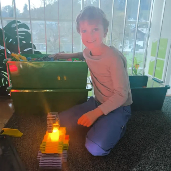 Ein junger Junge sitzt auf einem Teppichboden und spielt mit einem beleuchteten Spielzeugbagger. In der Nähe befinden sich grüne Aufbewahrungsboxen und ein Topf mit einer Pflanze ist draußen am Fenster zu sehen.