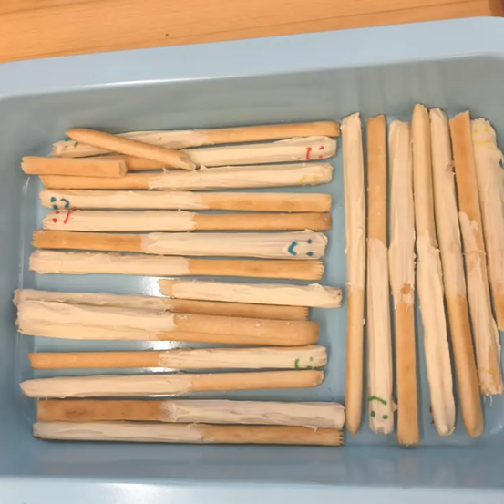 Eine Schale enthält zahlreiche Holzstäbchen, einige sind vollständig mit weißer Creme überzogen, andere nur teilweise. Einige Stäbchen haben gezeichnete Zahlen oder Symbole.