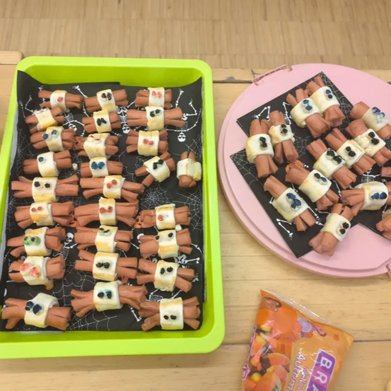 Zwei Tabletts mit Mini-Hot-Dog-Mumien auf schwarzen Tellern. Ein Tablett ist in einem grünen Behälter und das andere auf einem rosa Teller. Eine Tüte Süßigkeiten steht neben dem rosa Teller.