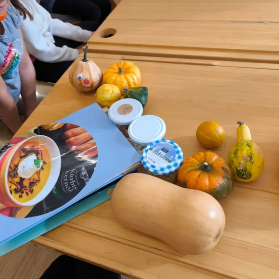Ein Holztisch zeigt verschiedene Kürbisse, Squashes und Lebensmittel, einschließlich eines pumkinsförmigen Buches und Behältern mit Deckeln. Ein Kind sitzt in der Nähe.