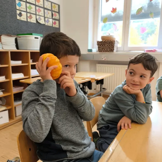 Zwei Jungen in einem Klassenzimmer, einer hält eine Kürbis und der andere stützt sein Kinn auf seine Hand. Dahinter eine Tafel mit 'Mit Brauner Eule' und Herbstblättern.