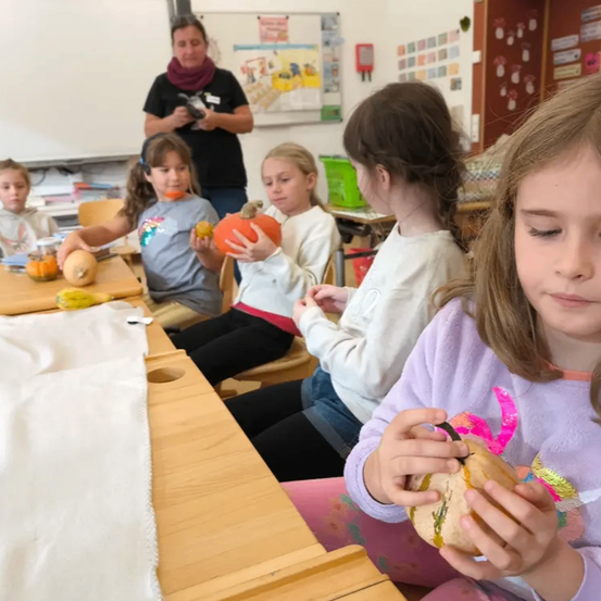 Eine Gruppe junger Mädchen in einem Klassenzimmer schnitzt Kürbisse, während ein Erwachsener beaufsichtigt. Jedes Mädchen hält einen Kürbis, und einige essen Kürbiskern. Das Klassenzimmer hat weiße Wände und eine Tafel.