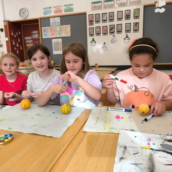 Vier Mädchen sitzen an einem Tisch in einem Klassenzimmer, jedes hält einen Marker. Sie malen gelbe Objekte, möglicherweise Kürbisse. Der Tisch ist mit Schutzfolien abgedeckt. Ein Bulletin-Board mit verschiedenen Bildern und Wörtern steht hinter ihnen.