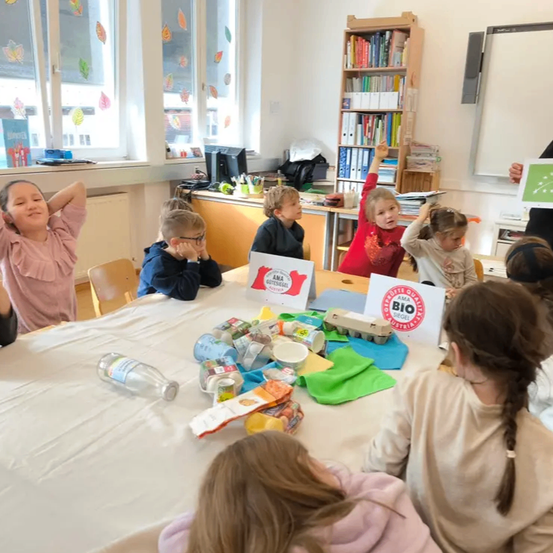 Kinder sitzen mit Essen und Getränken am Tisch, einige mit erhobenen Händen. Eines hält ein Schild für AMA Bio Siegel. Eine Frau steht in der Nähe.
