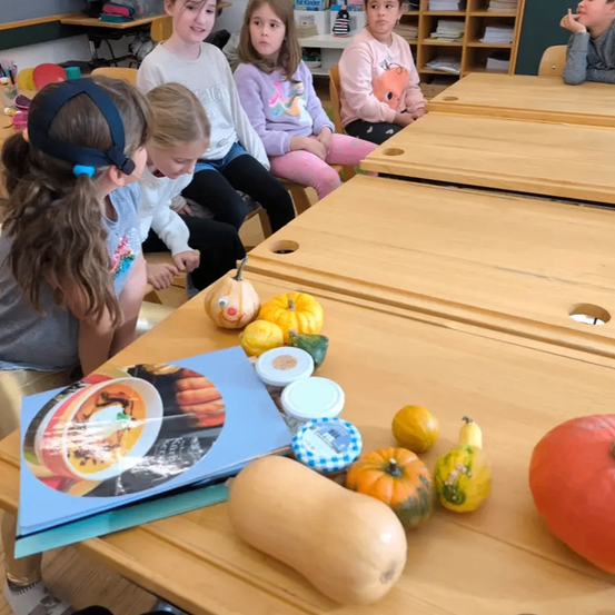 Eine Gruppe junger Mädchen sitzt um einen Holztisch in einem Klassenzimmer und betrachtet Kürbisse und ein Kochbuch.