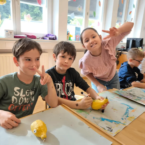 In einem Klassenzimmer sitzen drei Kinder um einen Tisch mit bemalten Papieren. Ein Junge in einem grünen Shirt hält ein gelbes Objekt, während ein Junge in einem schwarzen Shirt auf eine gelbe Banane malt. Ein Mädchen in einem rosa Shirt steht hinter ihnen mit erhobenen Armen.