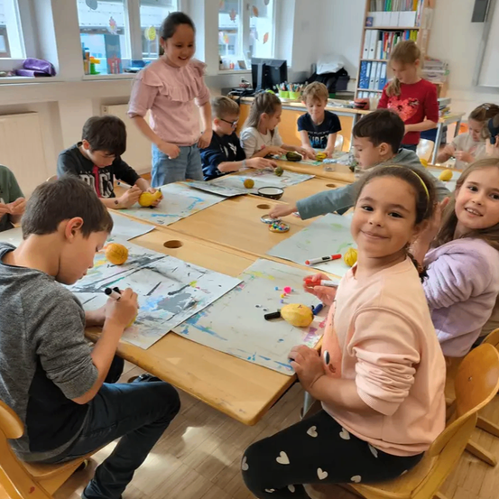 Kinder in einem Klassenzimmer arbeiten an Bastelarbeiten, einige mit Markern, während andere Zitronen als Requisiten verwenden. Ein Mädchen in Pink steht hinter ihnen und beobachtet.