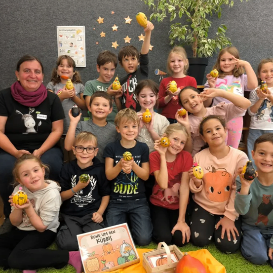 Eine Gruppe von Kindern und einem Erwachsenen posiert für ein Foto. Sie halten Kürbisse und lächeln. Der Erwachsene trägt eine Schürze.